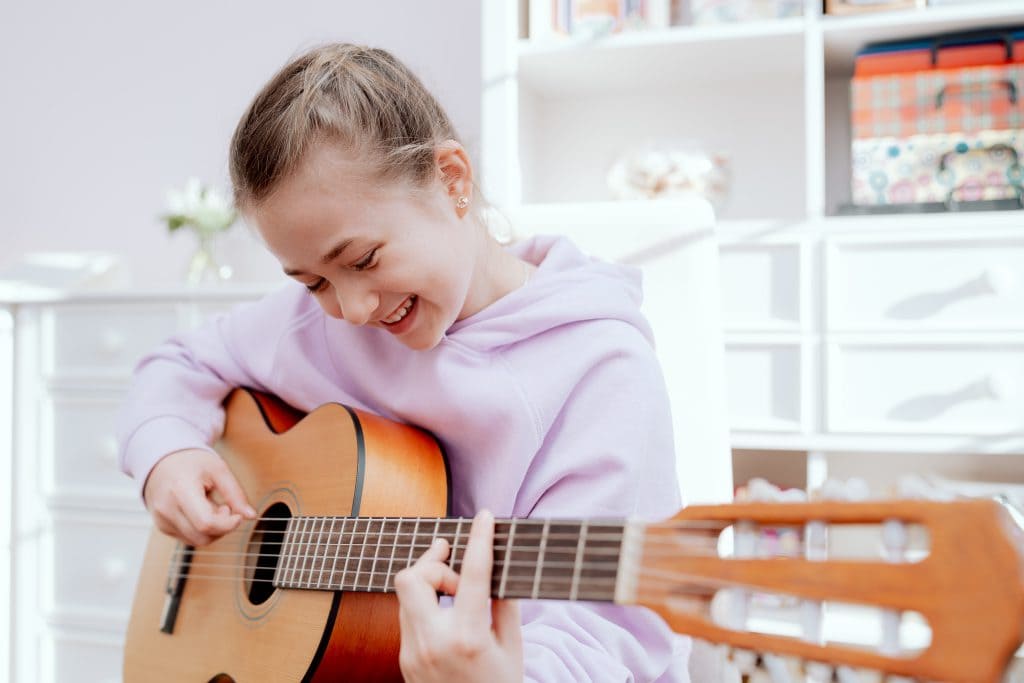 Smiling Caucasian teenage girl enjoys her free time playing acoustic guitar while sitting on a chair in her bright room.