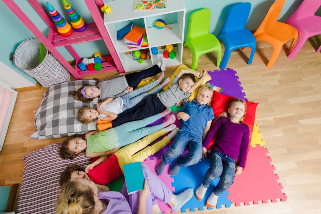 Young attractive teacher reading story before the naptime at the kindergarten. Lovely kids laying on a floor on soft mats and pillows. Time to have a rest and to calm down. Top view.