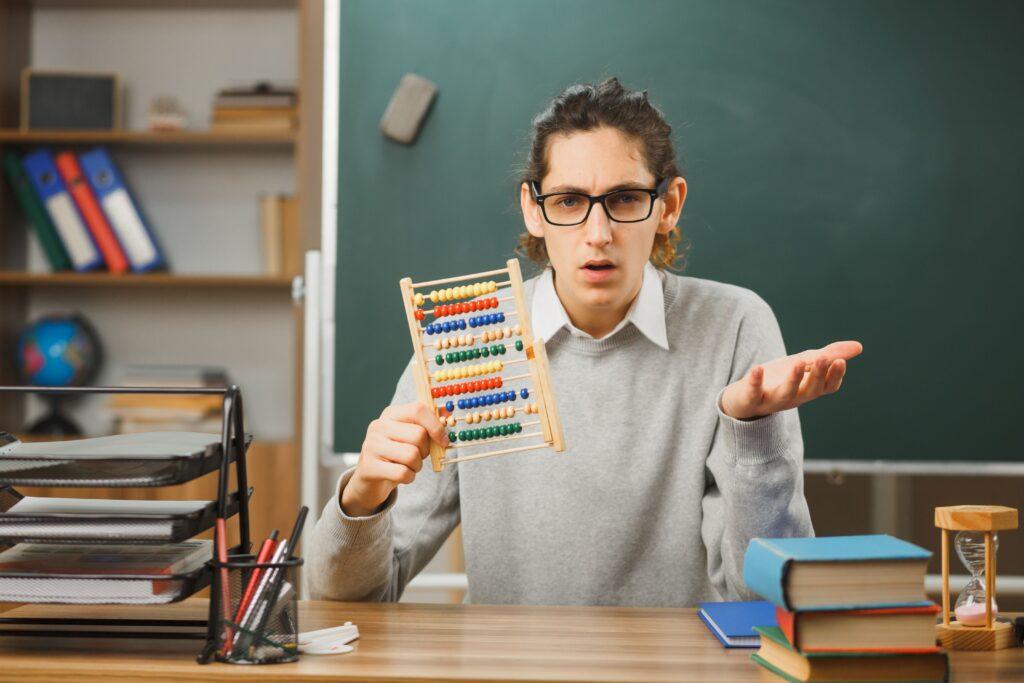 thinking young male teacher wearing glasses sitting desk holding abacus spread hands with school tools classroom